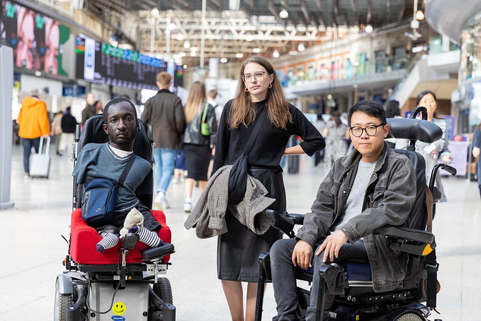 Three disabled people in a train station looking defiantly into the camera. On the left is a black man with upper and lower limb differences who uses an electric wheelchair. He has short brown hair and a beard, and is wearing a blue t-shirt and bag. In the middle is a white woman standing with her arm on her hip. She has brown hair, wears glasses, and a black jumper. On the right is an Asian man who uses an electric wheelchair. He has dark hair, and wears glasses, a grey denim jacket, and jeans.